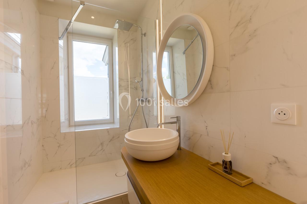 Salle de bain moderne avec lavabo rond, miroir éclairé, douche vitrée et murs en carrelage effet marbre.