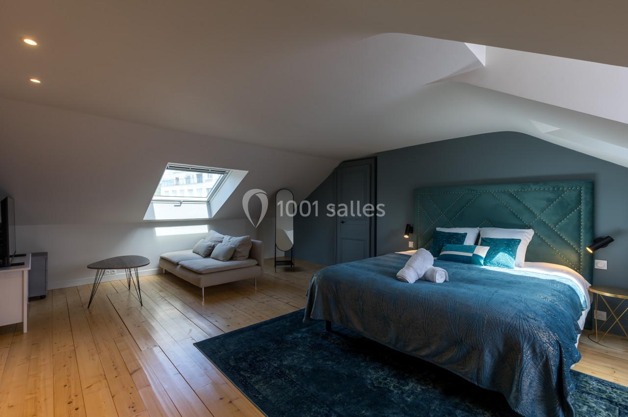 Chambre mansardée avec lit double, canapé, miroir sur pied, lumière naturelle et parquet en bois clair.