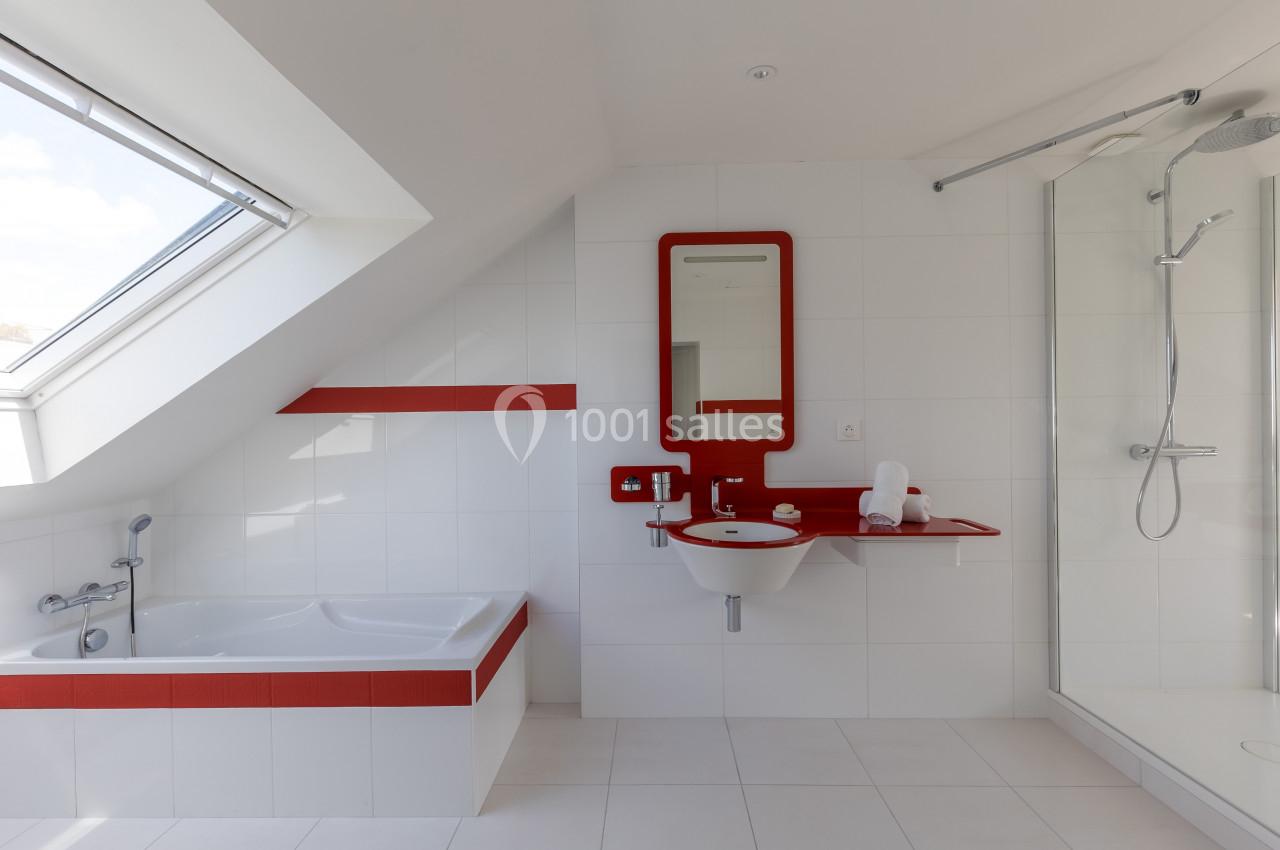 Salle de bain moderne avec baignoire, douche, lavabo et miroir encadrés de rouge, sous un toit mansardé avec fenêtre.