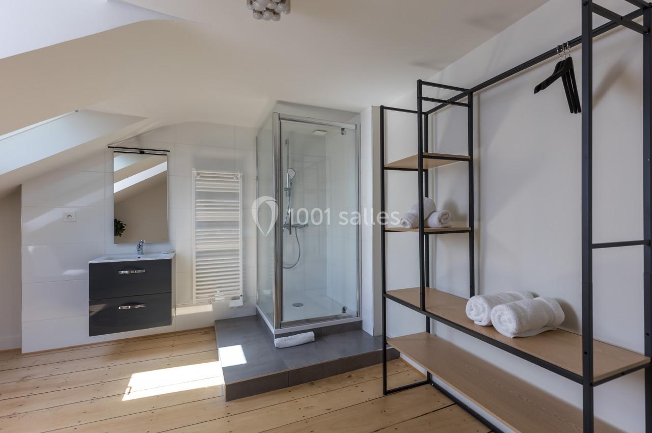Salle de bain mansardée avec douche vitrée, meuble lavabo, étagères en métal et bois, et serviettes roulées.