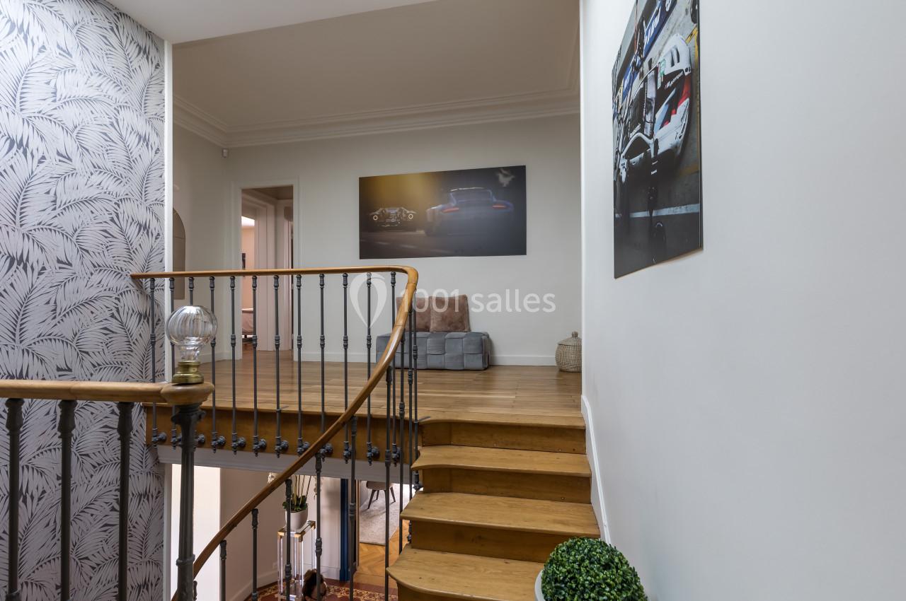 Escalier en bois avec rampe en métal, menant à un palier décoré de tableaux et d'un canapé gris.