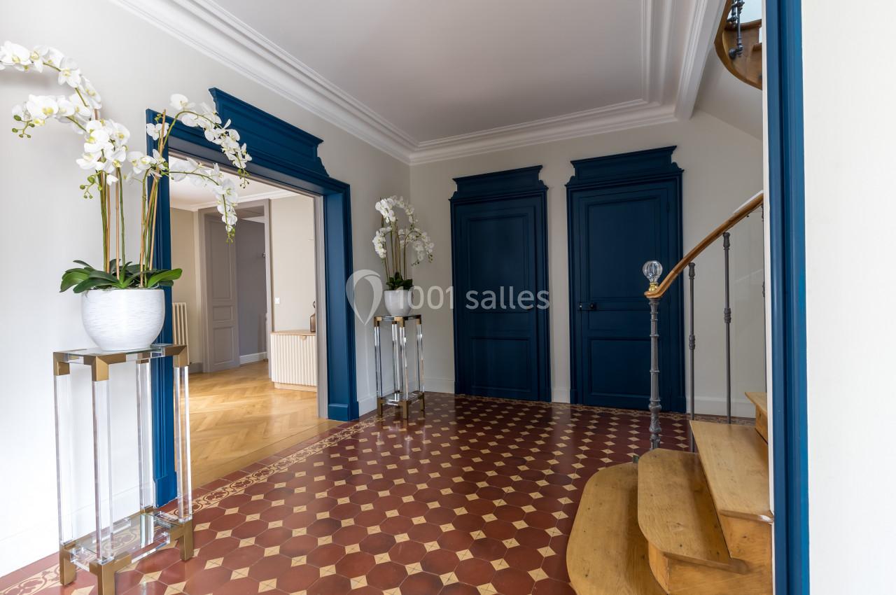 Entrée lumineuse avec carrelage à motifs, murs blancs, portes bleues, escaliers en bois et orchidées en décoration.