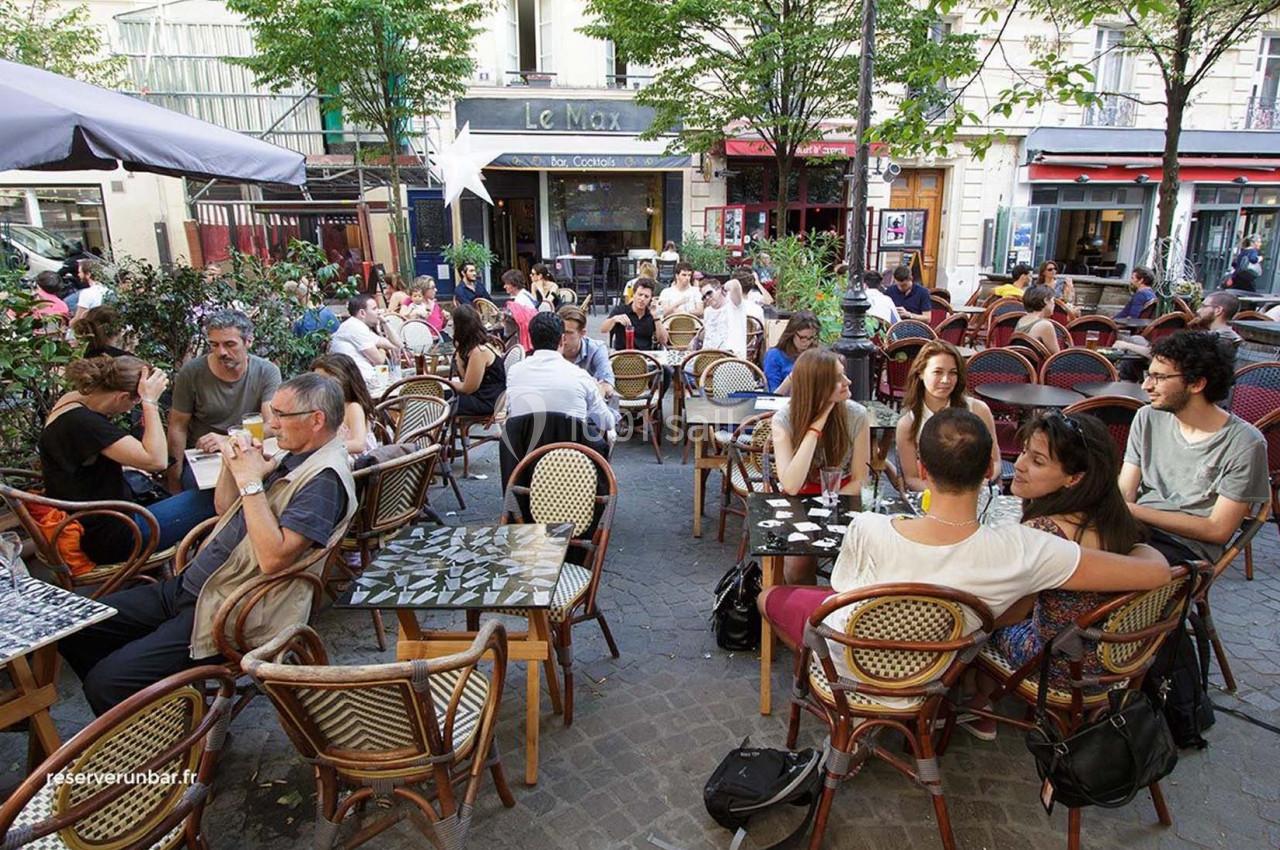 Terrasse animée d'un café avec des clients assis à des tables en plein air, entourée d'arbres et de bâtiments.