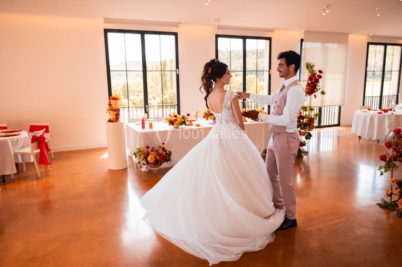 Un couple danse dans une salle lumineuse décorée de fleurs et de tables pour une réception.
