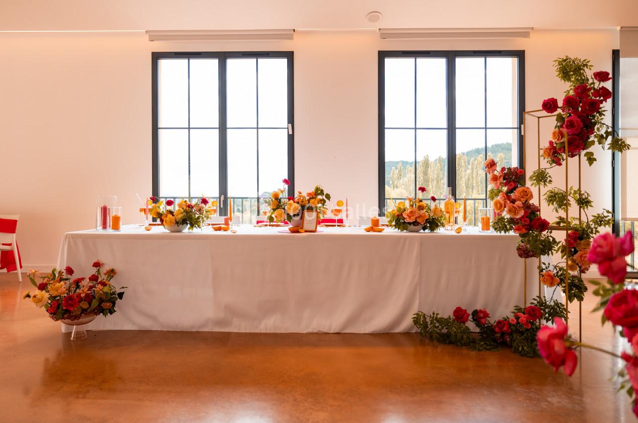Table décorée avec des fleurs colorées, des bougies et des éléments de vaisselle, placée devant de grandes fenêtres.