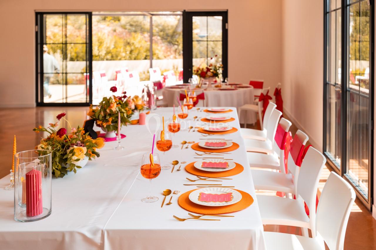 Table dressée avec des assiettes colorées, verres orange et décorations florales dans une salle lumineuse avec grandes baies…