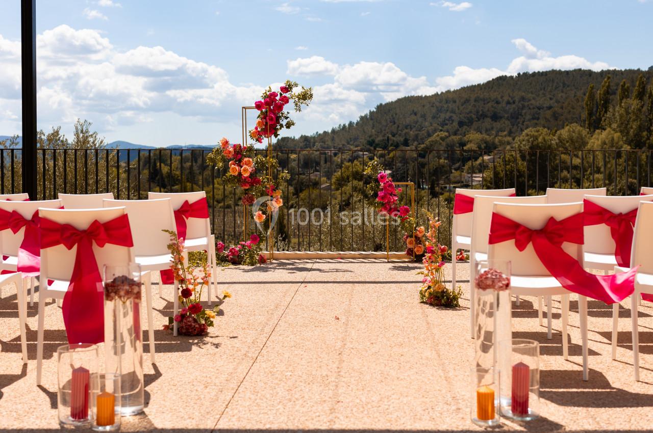 Chaises blanches décorées de nœuds rouges face à une arche florale colorée en extérieur, avec vue sur des collines boisées.