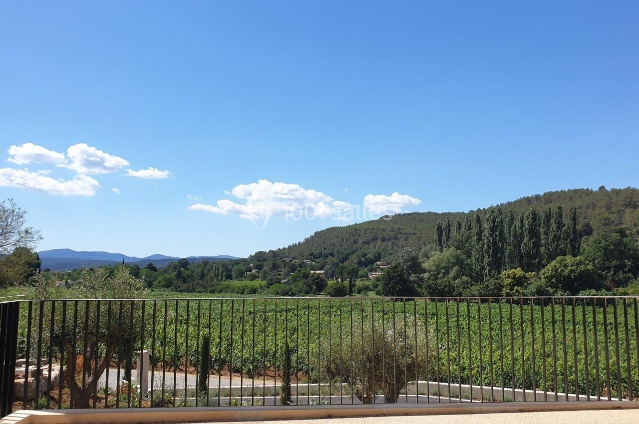 Vue sur un paysage rural avec des collines boisées, des vignes et un ciel bleu, depuis une terrasse avec une barrière noire.