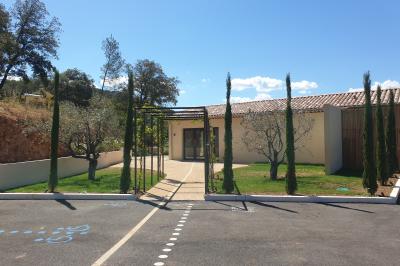 Entrée d'un bâtiment moderne entourée d'arbres et de cyprès, avec une allée pavée et un ciel dégagé en arrière-plan.