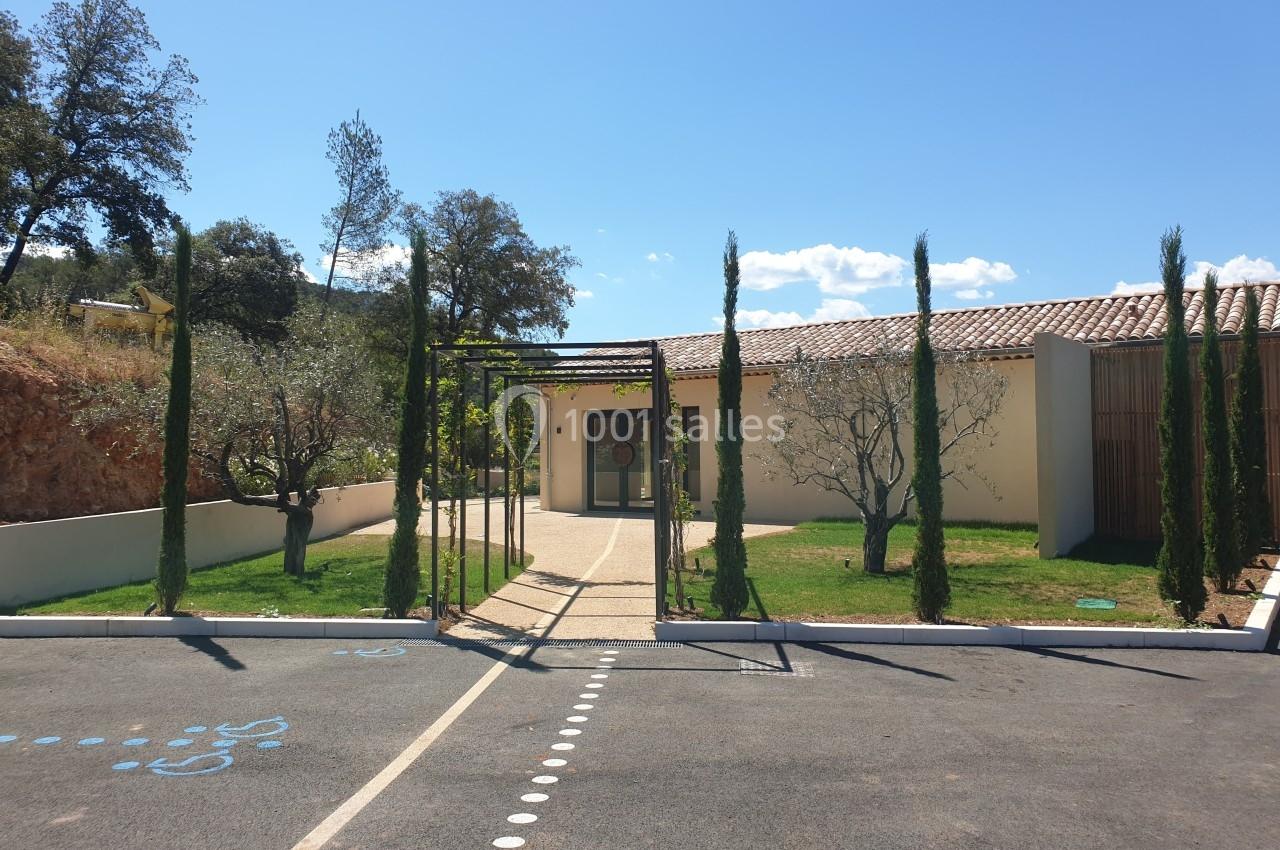 Entrée d'un bâtiment moderne entourée d'arbres et de cyprès, avec une allée pavée et un ciel dégagé en arrière-plan.