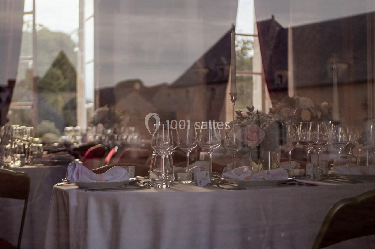 Salle de réception avec tables dressées, nappes blanches, verres et décorations florales, vue à travers une vitre.