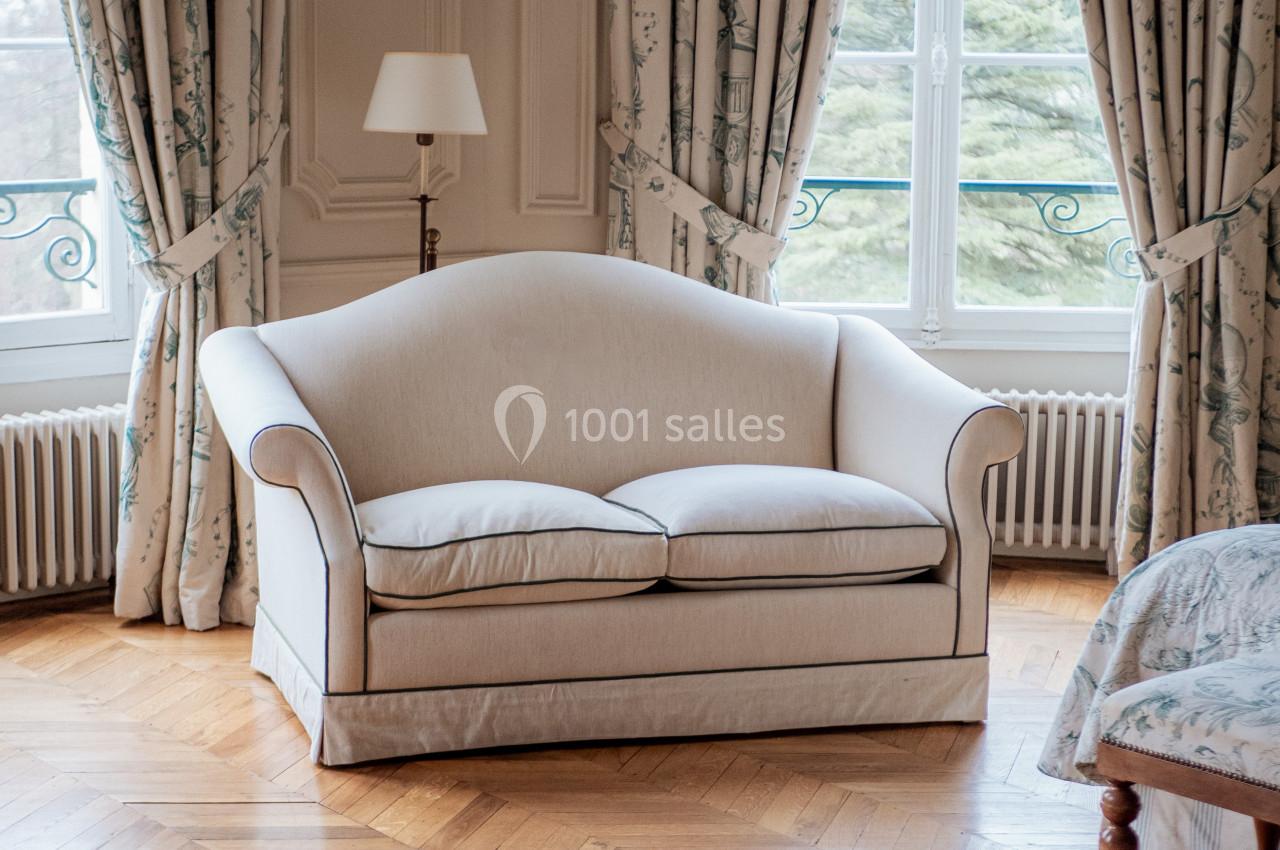 Canapé beige à deux places dans un salon lumineux avec parquet, rideaux à motifs et vue sur un jardin.