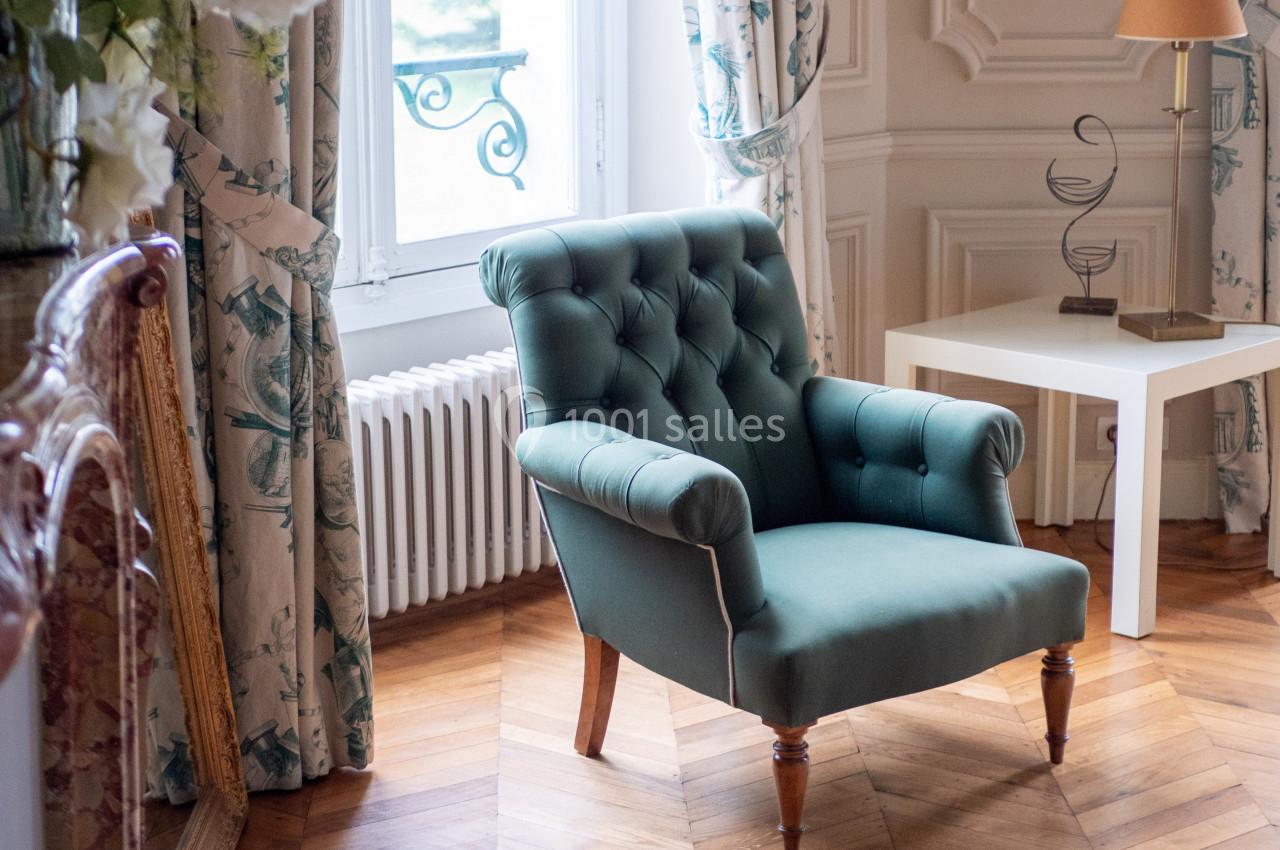 Fauteuil vert capitonné placé dans un salon lumineux avec parquet, près d'une fenêtre et d'une table blanche.
