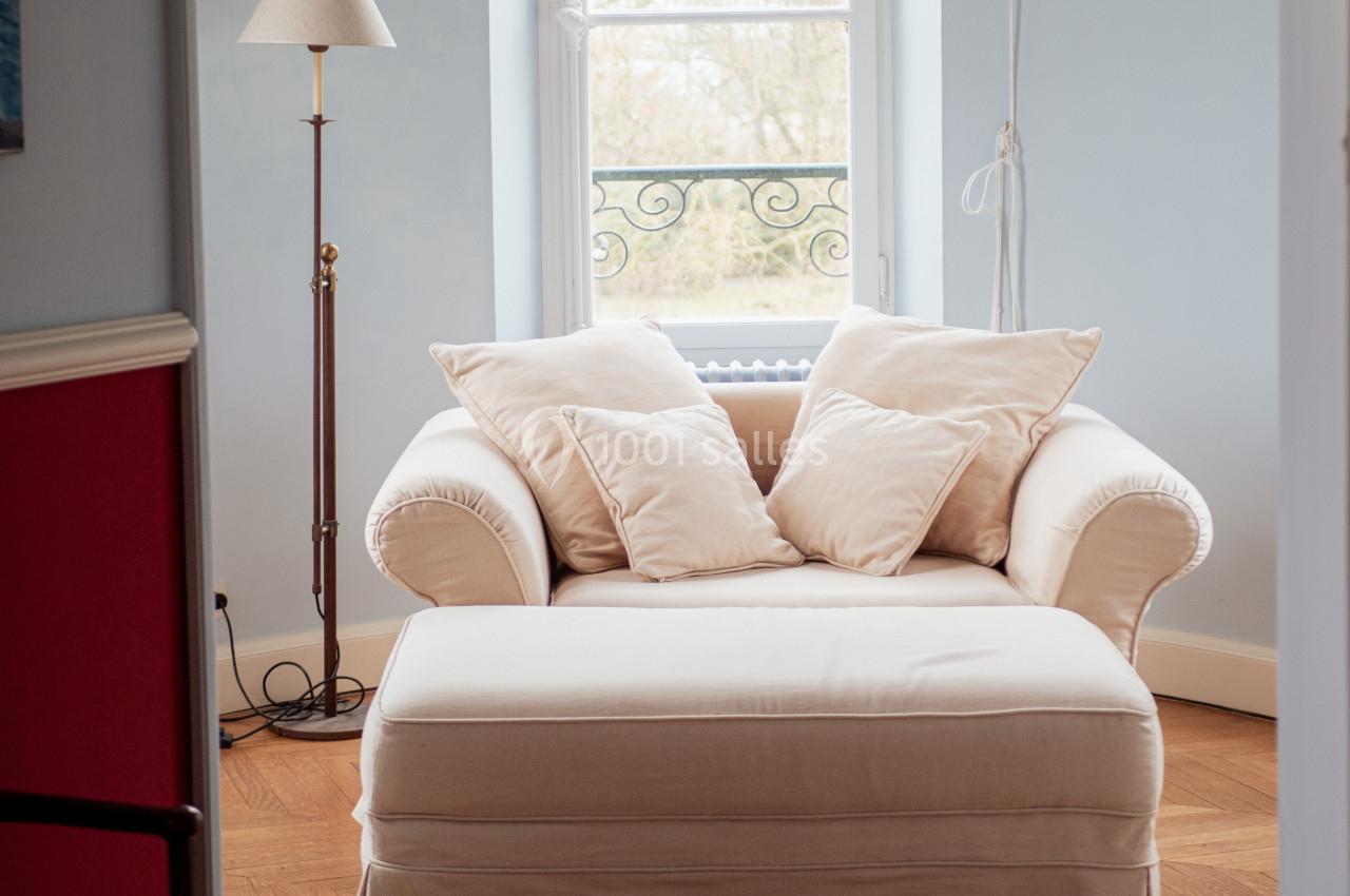 Fauteuil beige avec repose-pieds assorti, placé près d'une fenêtre lumineuse dans une pièce aux tons clairs.