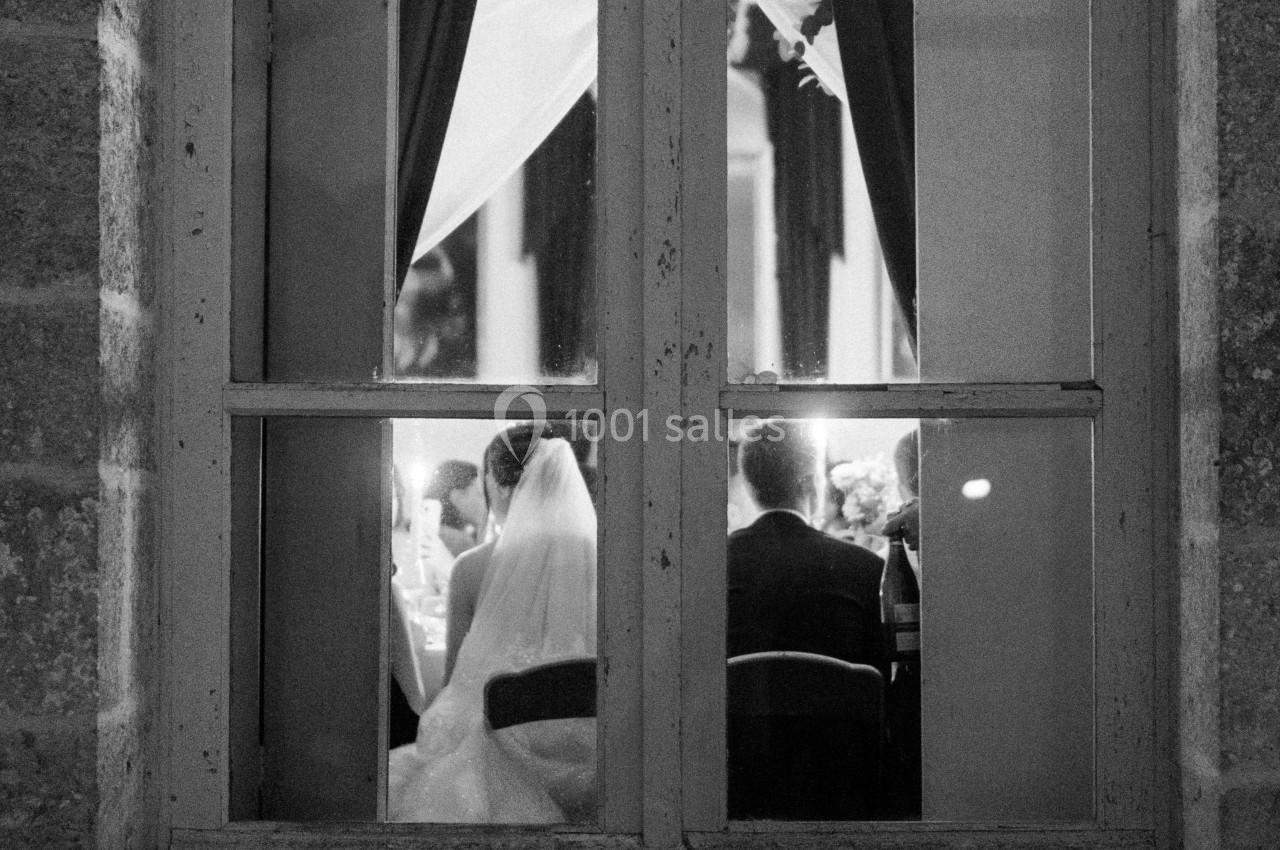 Vue à travers une fenêtre, un couple de mariés assis de dos lors d'une réception éclairée.