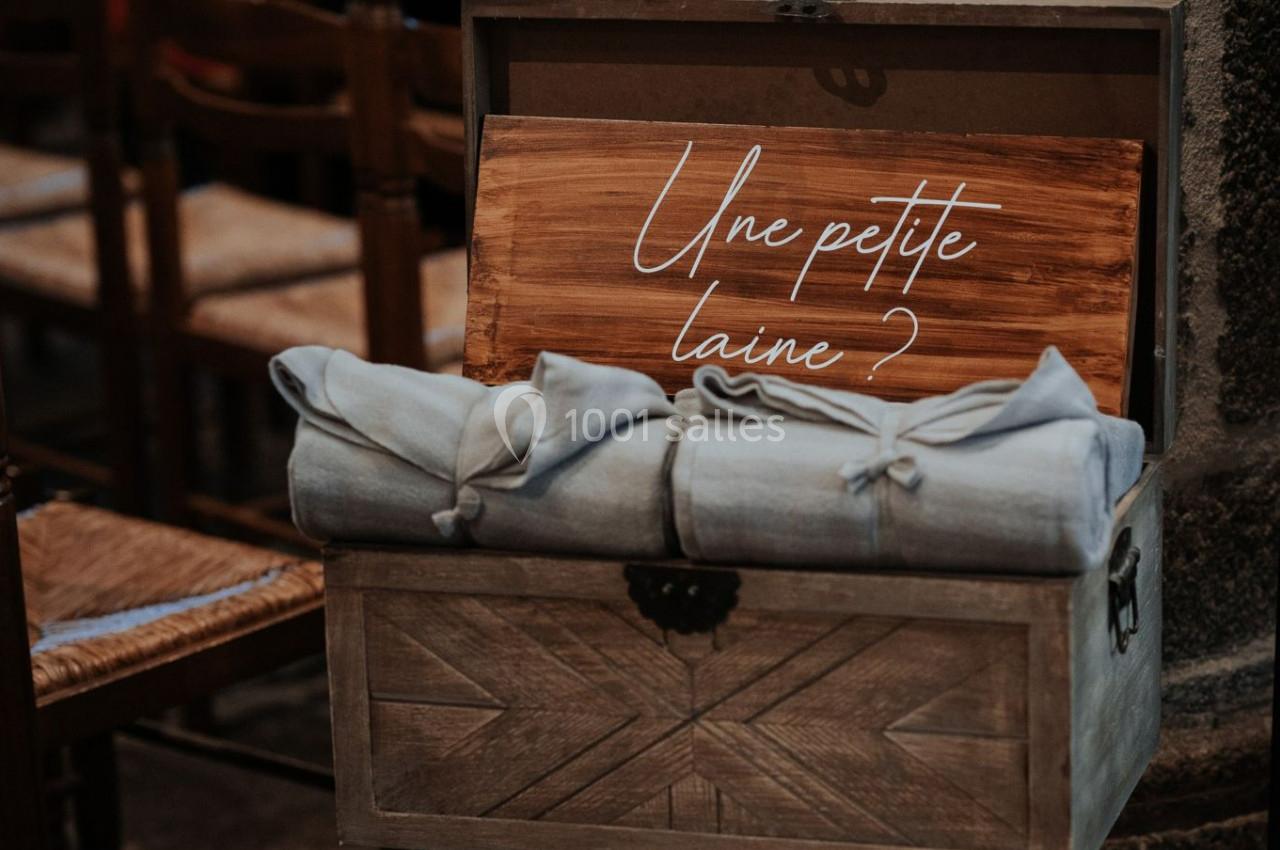 Coffre en bois ouvert contenant des couvertures pliées et une pancarte en bois avec l'inscription ’Une petite laine ?’.