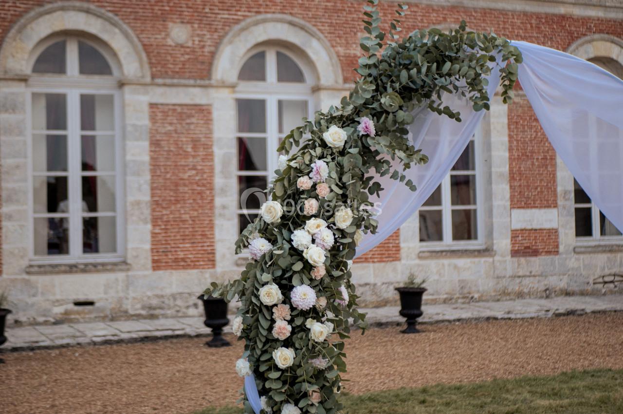 Arche décorée de fleurs blanches et roses avec feuillage, installée devant une façade en briques et fenêtres cintrées.