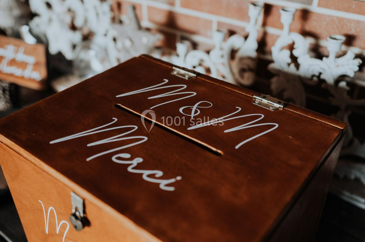 Boîte en bois avec une fente, décorée des inscriptions ’M & M’ et ’Merci’, posée devant un mur en briques.
