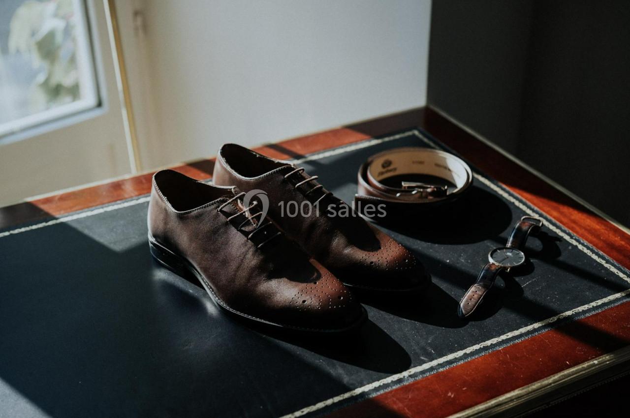 Chaussures en cuir marron avec ceinture assortie et montre posées sur un bureau en bois près d'une fenêtre.