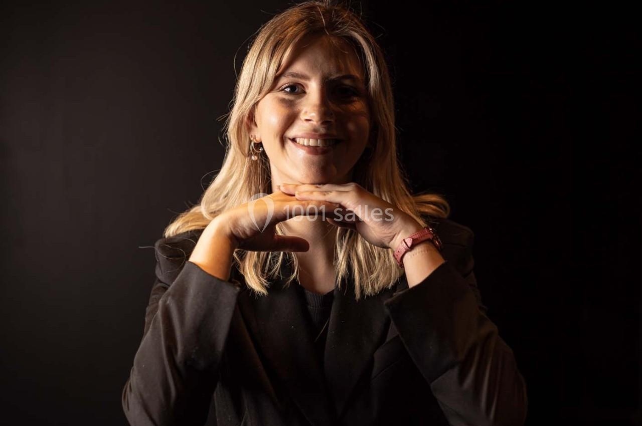 Femme souriante posant dans un environnement sombre, appuyée sur ses mains croisées sous son menton.