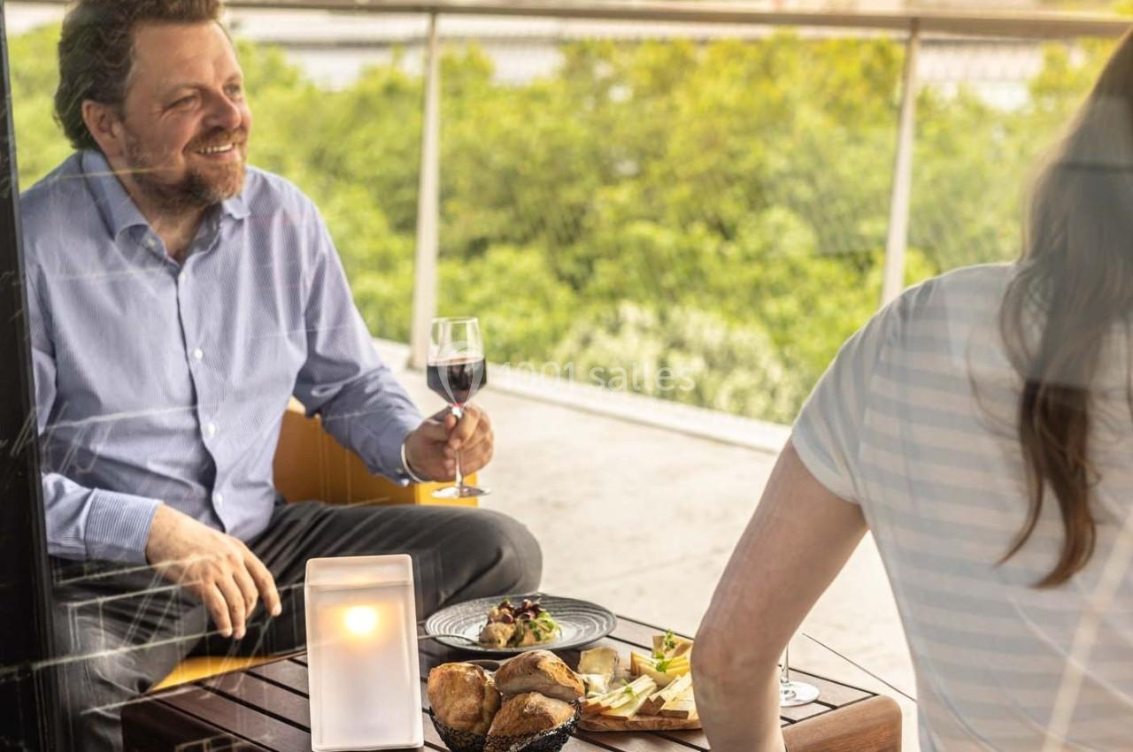 Un homme souriant tenant un verre de vin, assis à une table avec des plats, sur une terrasse avec vue sur la verdure.
