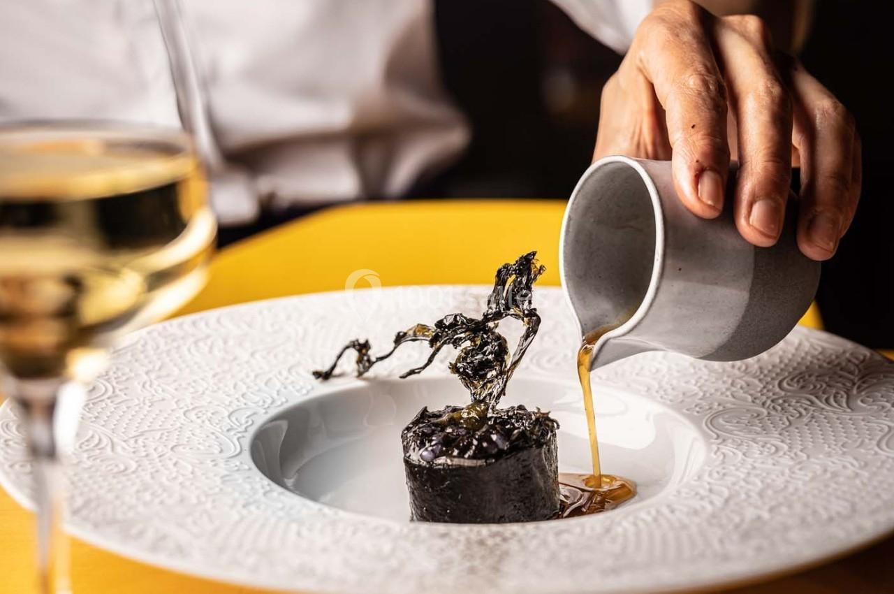 Une main verse une sauce dans une assiette contenant un plat raffiné décoré d'algues noires.