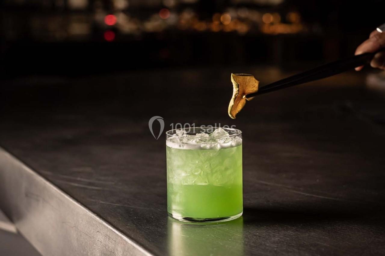 Verre de cocktail vert avec glaçons sur une table métallique, une main ajoutant une garniture dorée avec des pinces.
