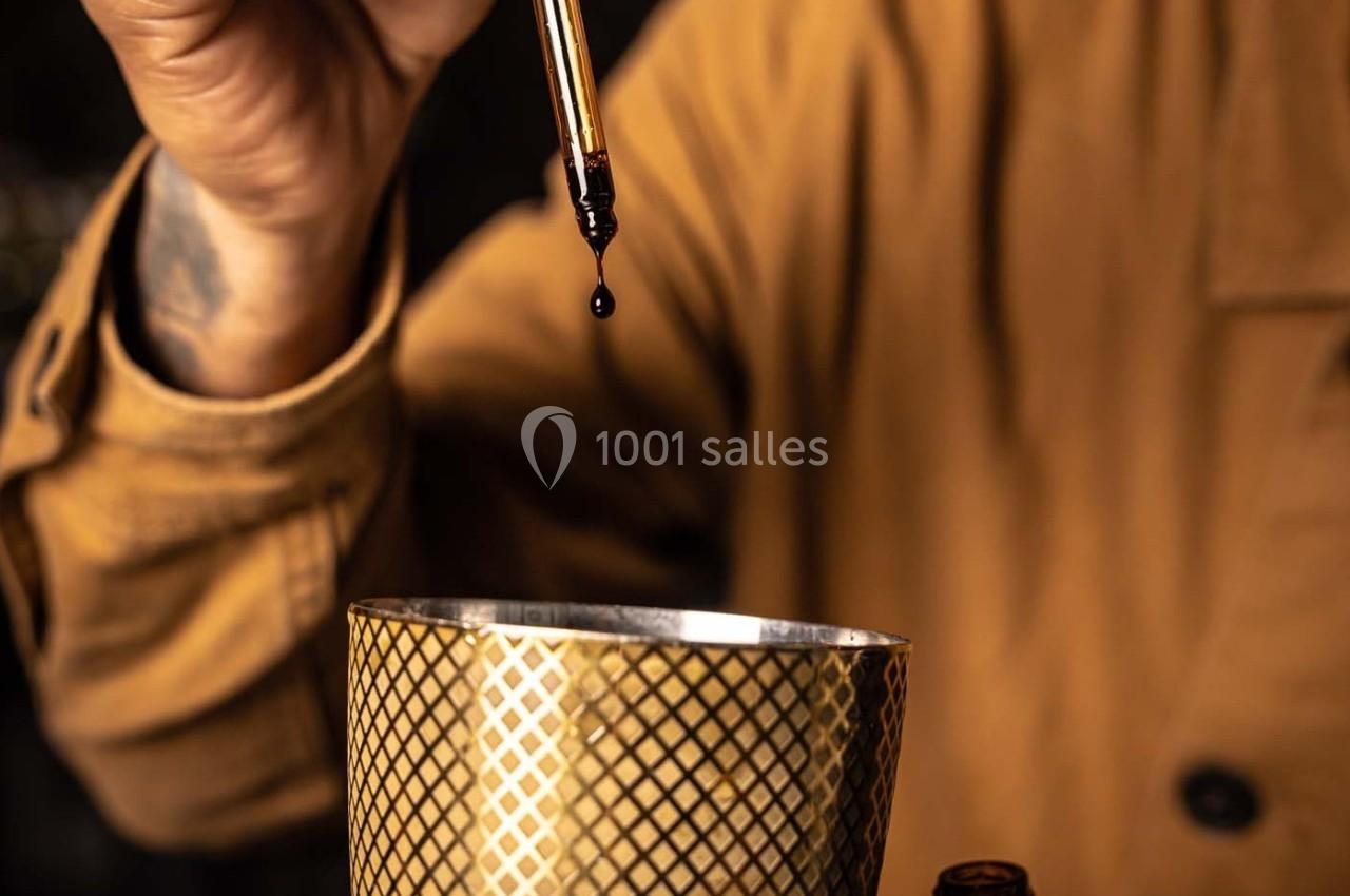 Une main tient une pipette laissant tomber une goutte de liquide dans un récipient doré à motifs géométriques.