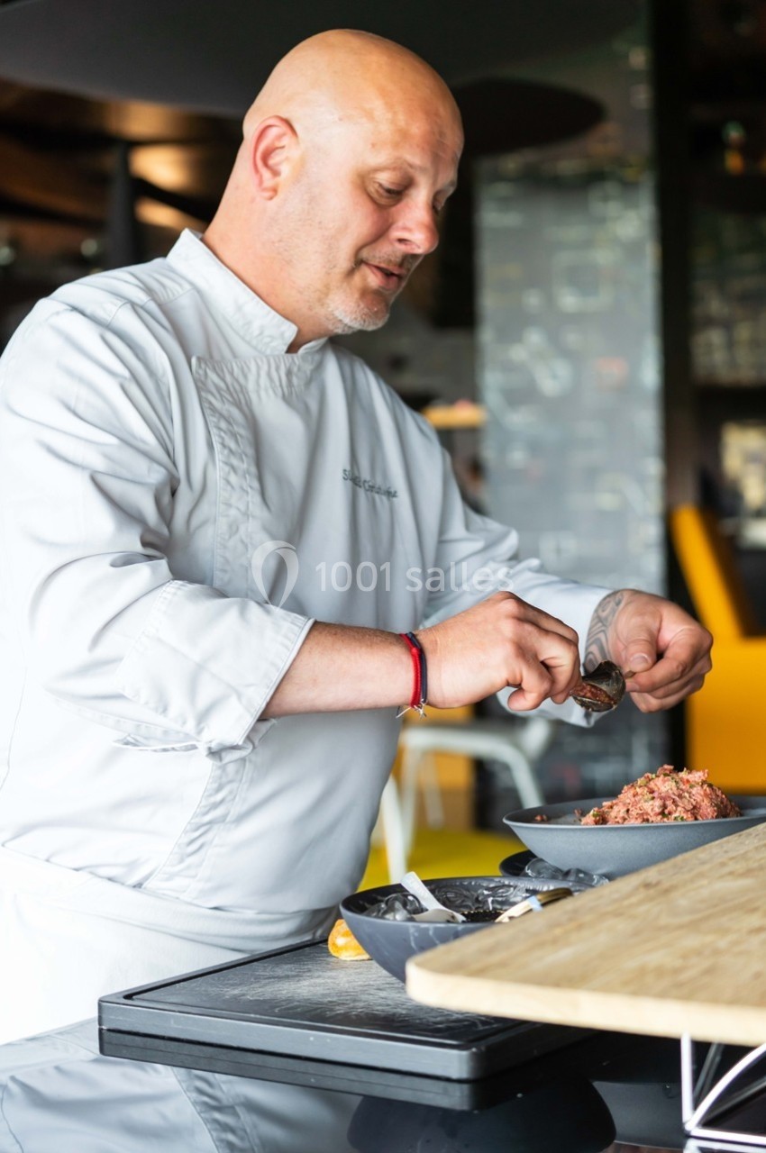 Un chef en tenue blanche prépare un plat en disposant des ingrédients dans un bol dans une cuisine professionnelle.