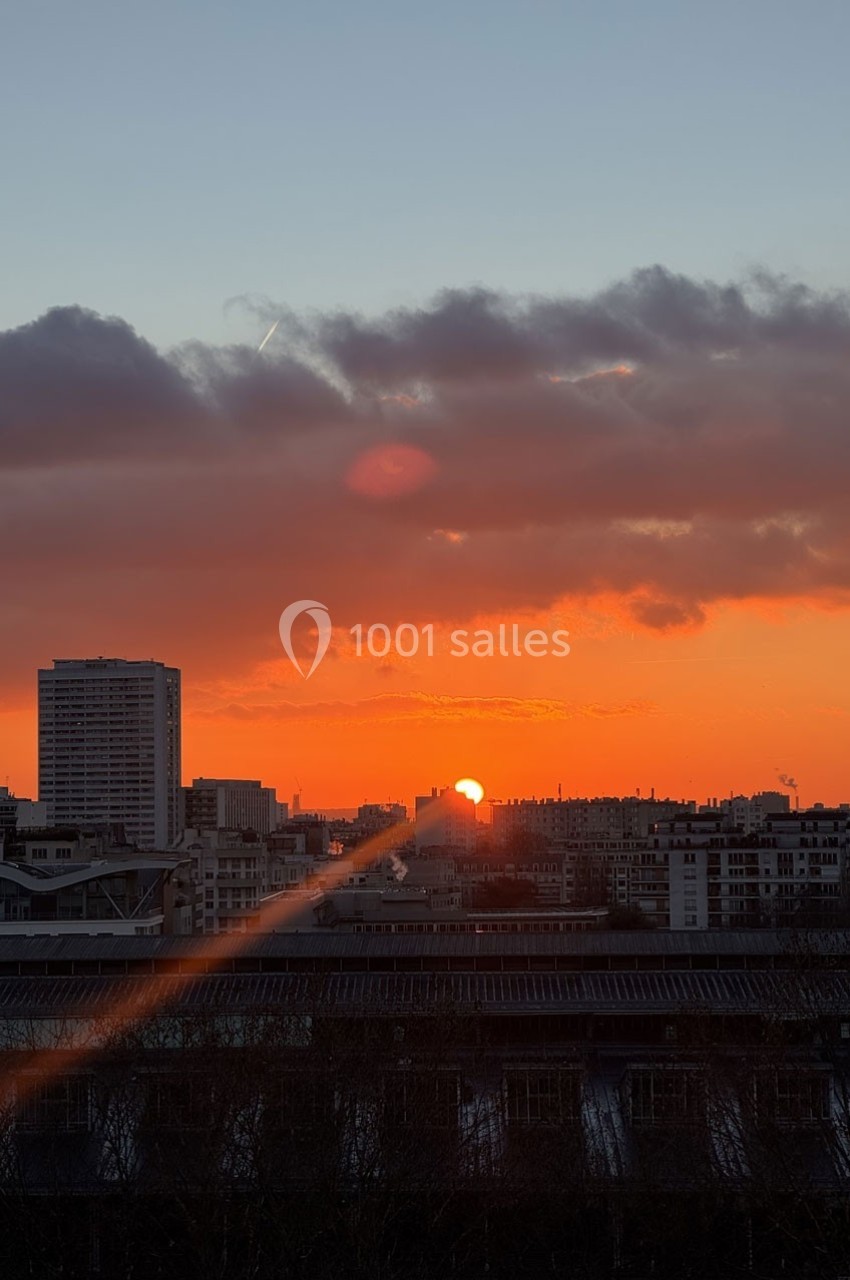 Coucher de soleil sur une ville avec des immeubles, ciel orangé et nuages sombres à l'horizon.