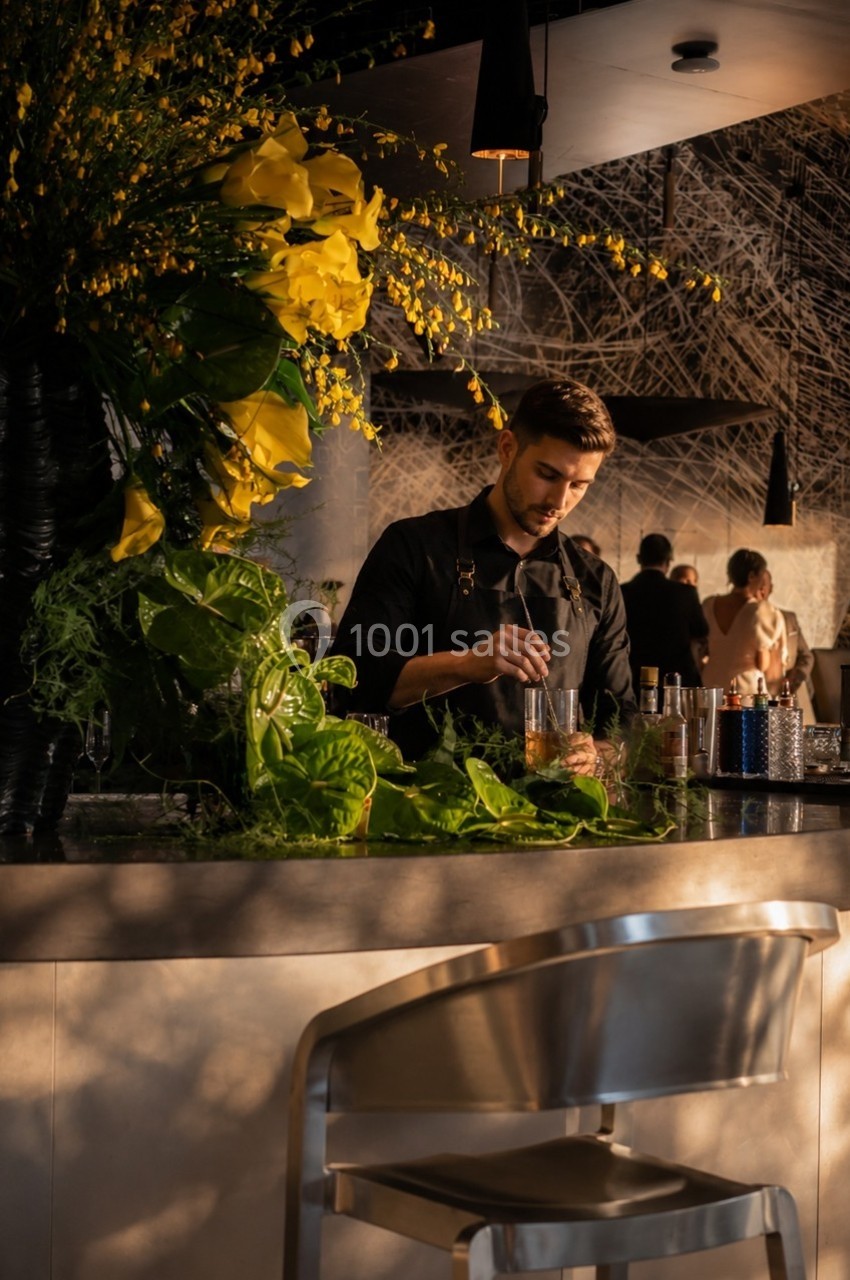 Un barman prépare un cocktail dans un bar élégant, décoré de grandes compositions florales jaunes.