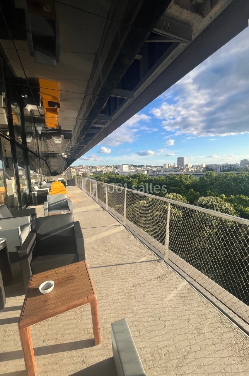Terrasse en hauteur avec mobilier moderne, vue dégagée sur une ville et un parc sous un ciel partiellement nuageux.