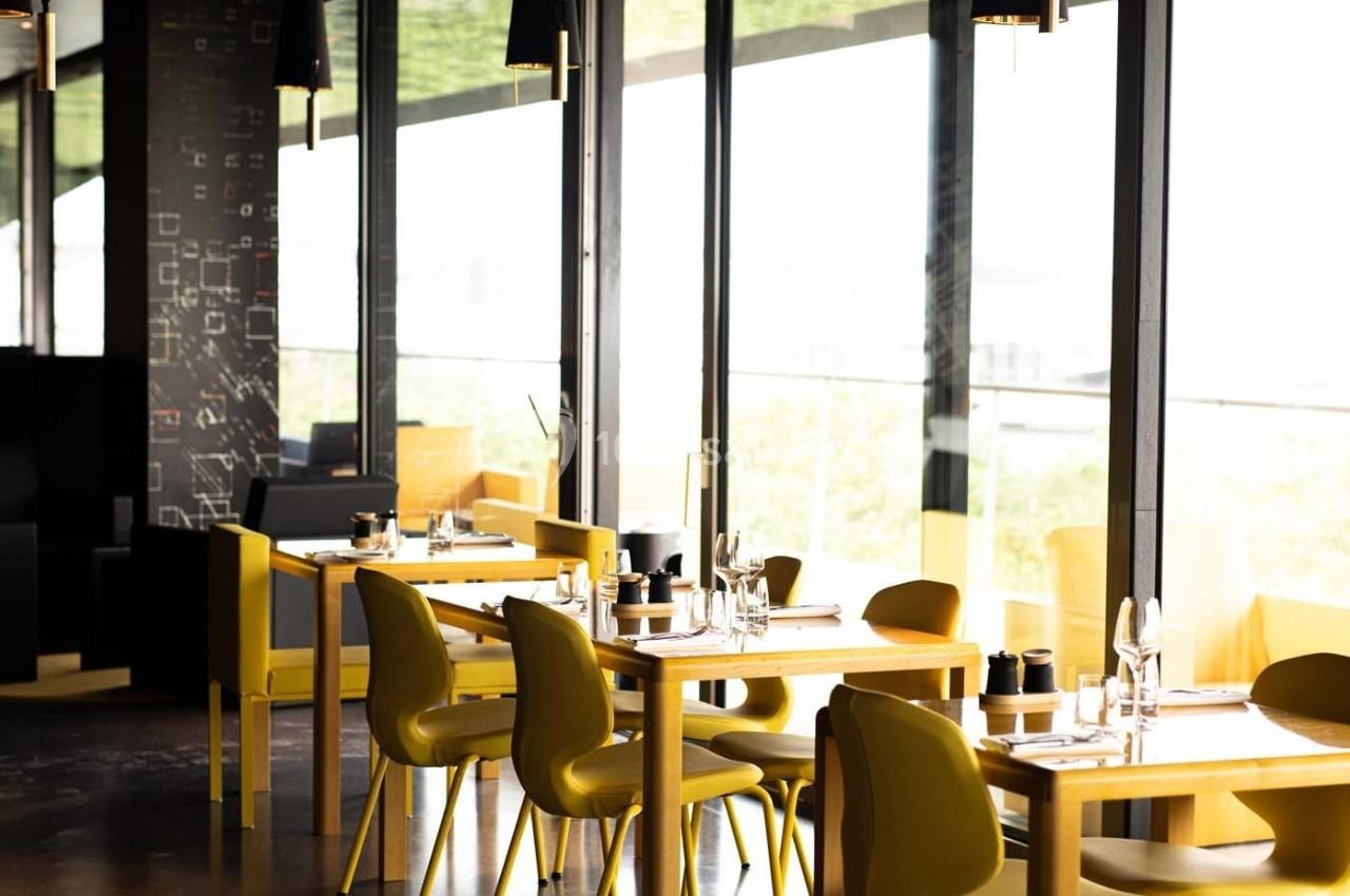 Salle de restaurant lumineuse avec des tables en bois clair, des chaises jaunes et de grandes baies vitrées.