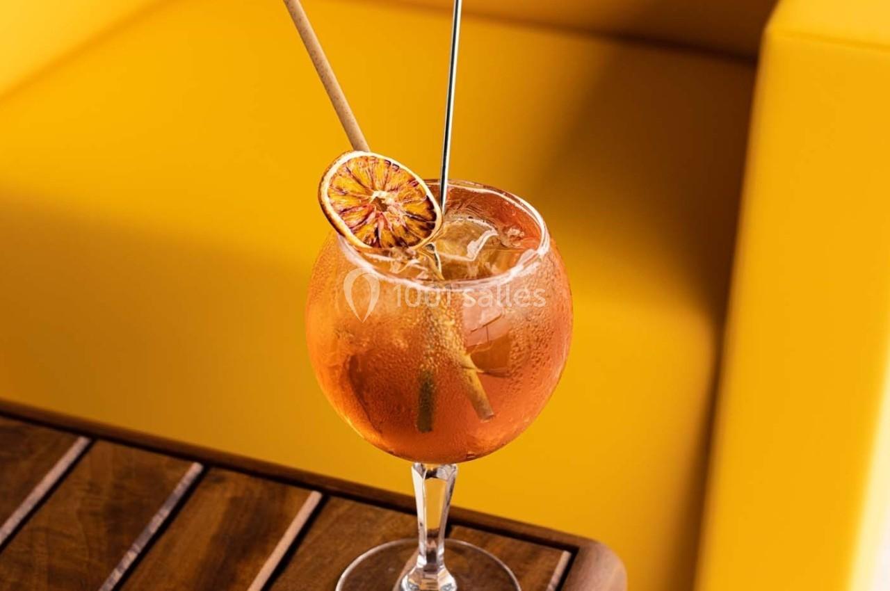 Verre de cocktail orange garni d'une tranche d'agrume séché, posé sur une table en bois devant un fond jaune.