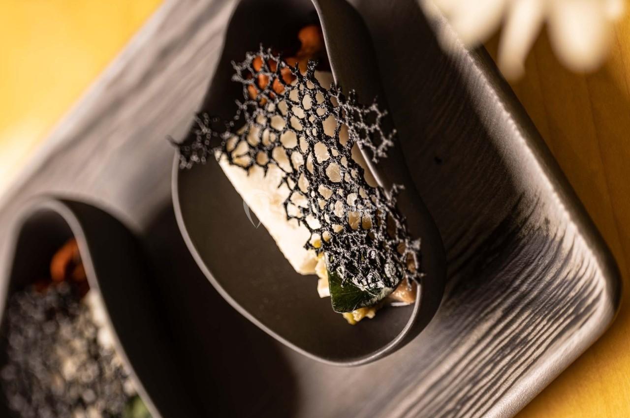 Plat gastronomique présenté dans une assiette noire, décoré d'une dentelle alimentaire noire et de légumes.