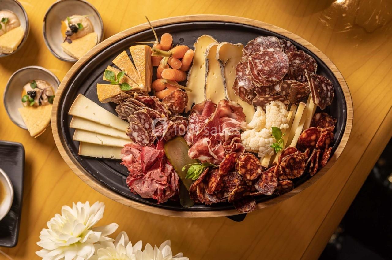 Plateau de charcuterie et fromages variés avec légumes marinés, présenté sur une table en bois clair.