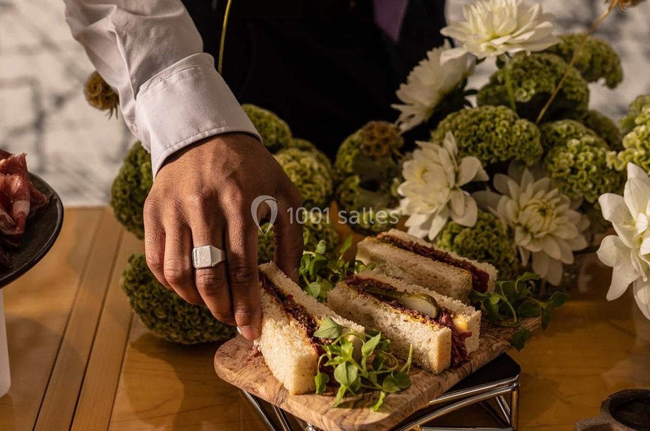 Une main saisit un sandwich garni sur une planche en bois, entourée de fleurs et de verdure décorative.