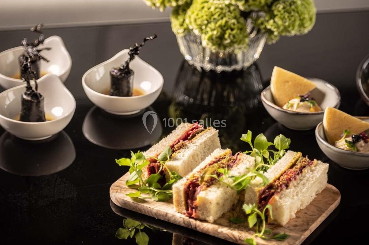 Assiette de sandwichs garnis de viande et légumes, entourée de petites entrées sur une table noire élégante.