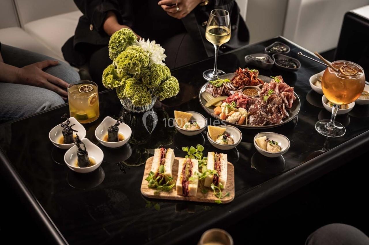 Table noire avec cocktails, assiettes de tapas variées, fleurs décoratives et convives en arrière-plan.