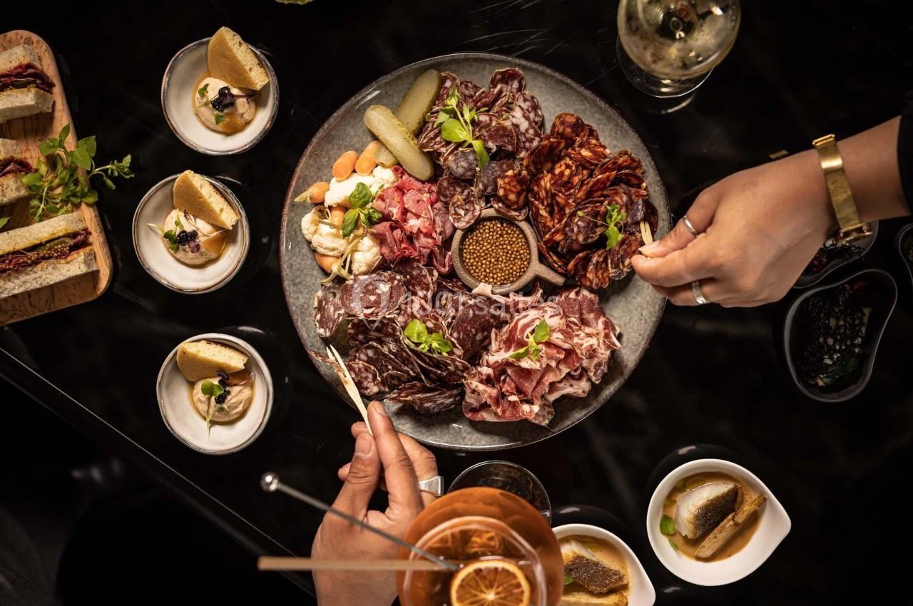 Plateau de charcuteries variées avec condiments, accompagné de petits plats et boissons sur une table sombre.