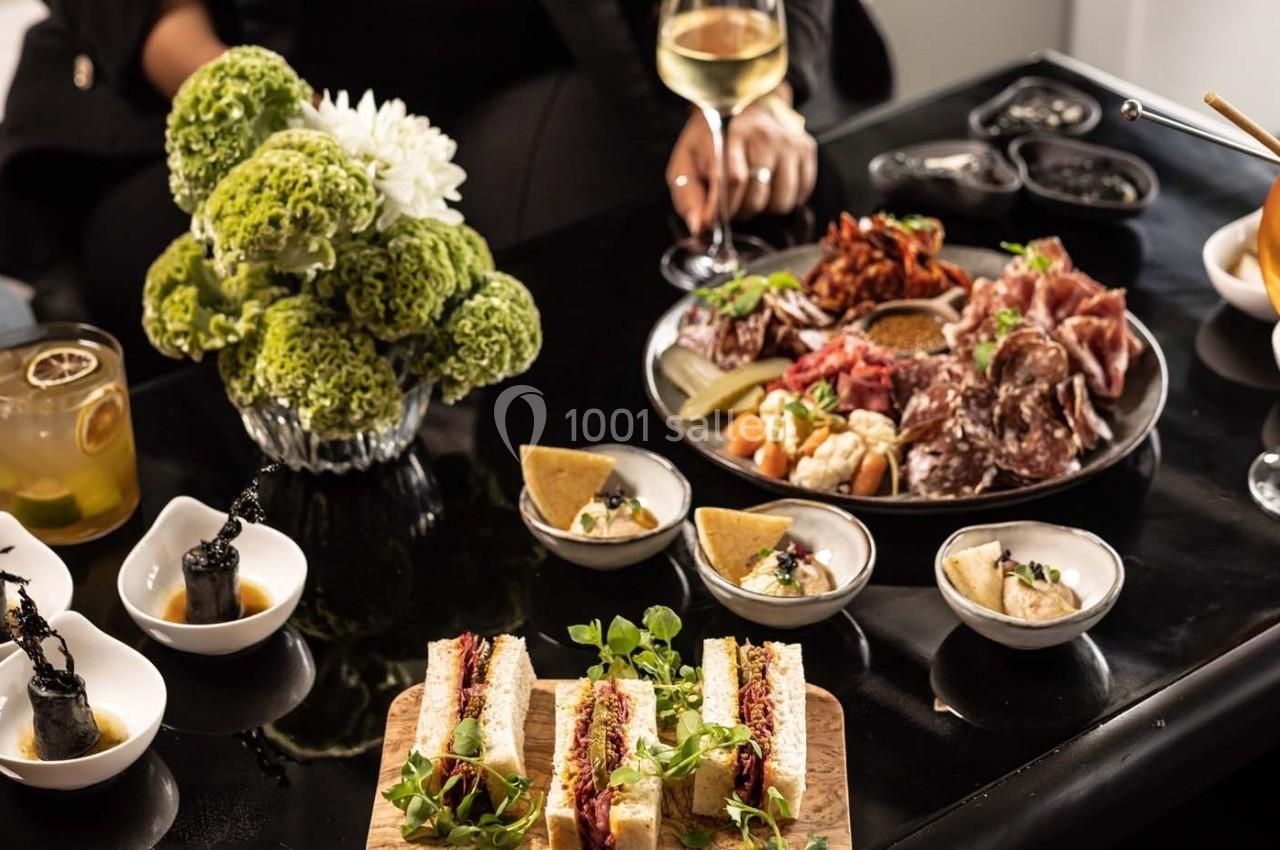 Plateau de charcuterie, amuse-bouches variés et sandwichs gourmands sur une table avec boissons et décoration florale.