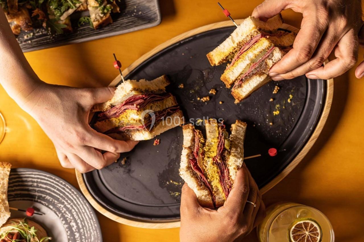 Trois mains tenant des sandwichs garnis de viande et fromage sur un plateau, avec des assiettes et un verre en arrière-plan.