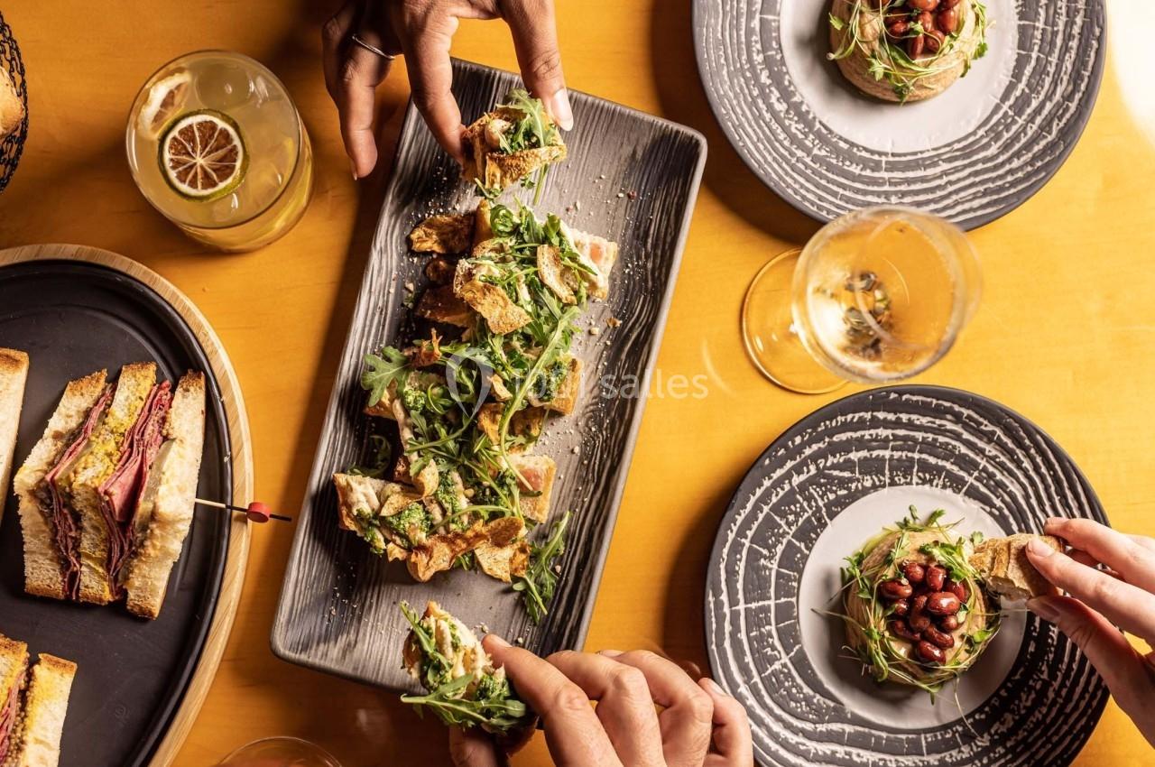 Des mains partagent des plats variés sur une table, comprenant des toasts garnis, un sandwich et des boissons.