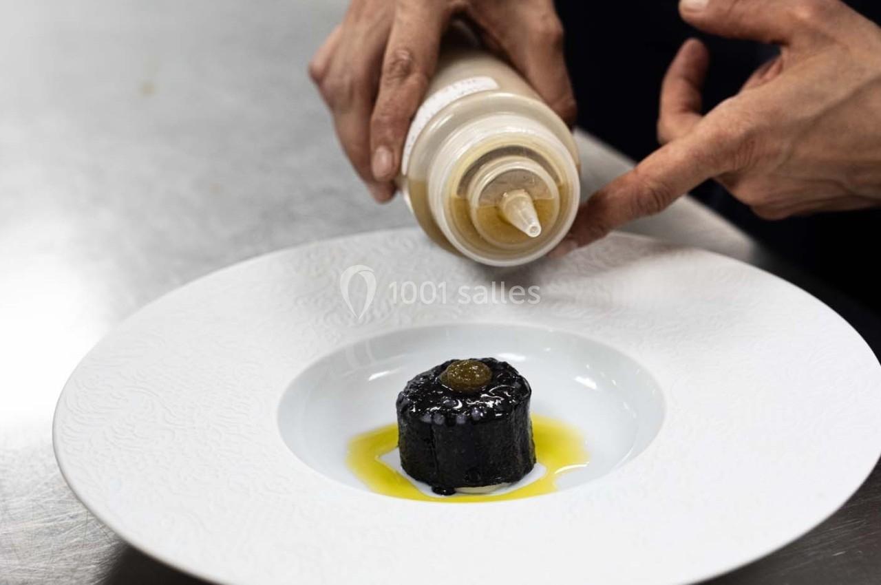 Un chef verse une sauce sur un plat raffiné composé d'un cylindre noir et d'une garniture dans une assiette blanche.