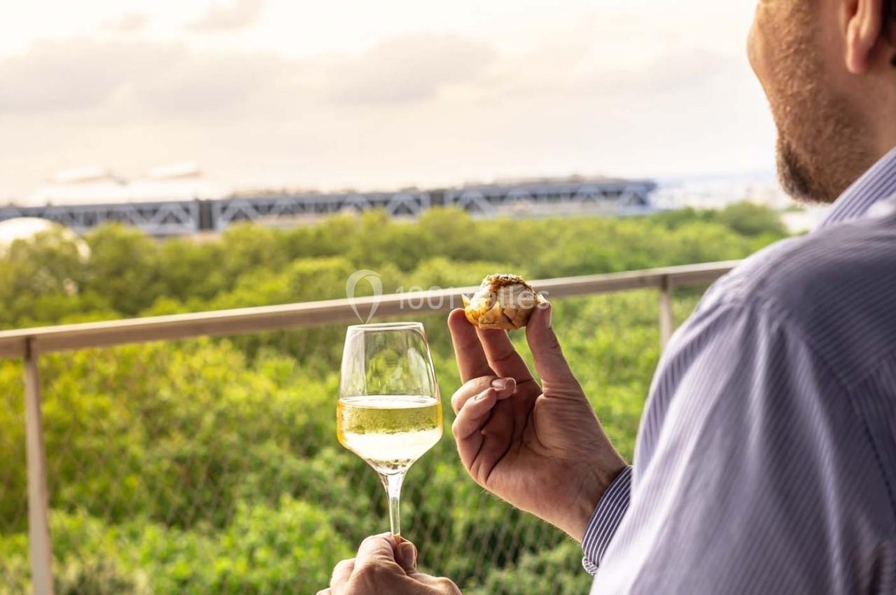 Un homme tient un verre de vin blanc et un amuse-bouche sur une terrasse surplombant un paysage verdoyant.