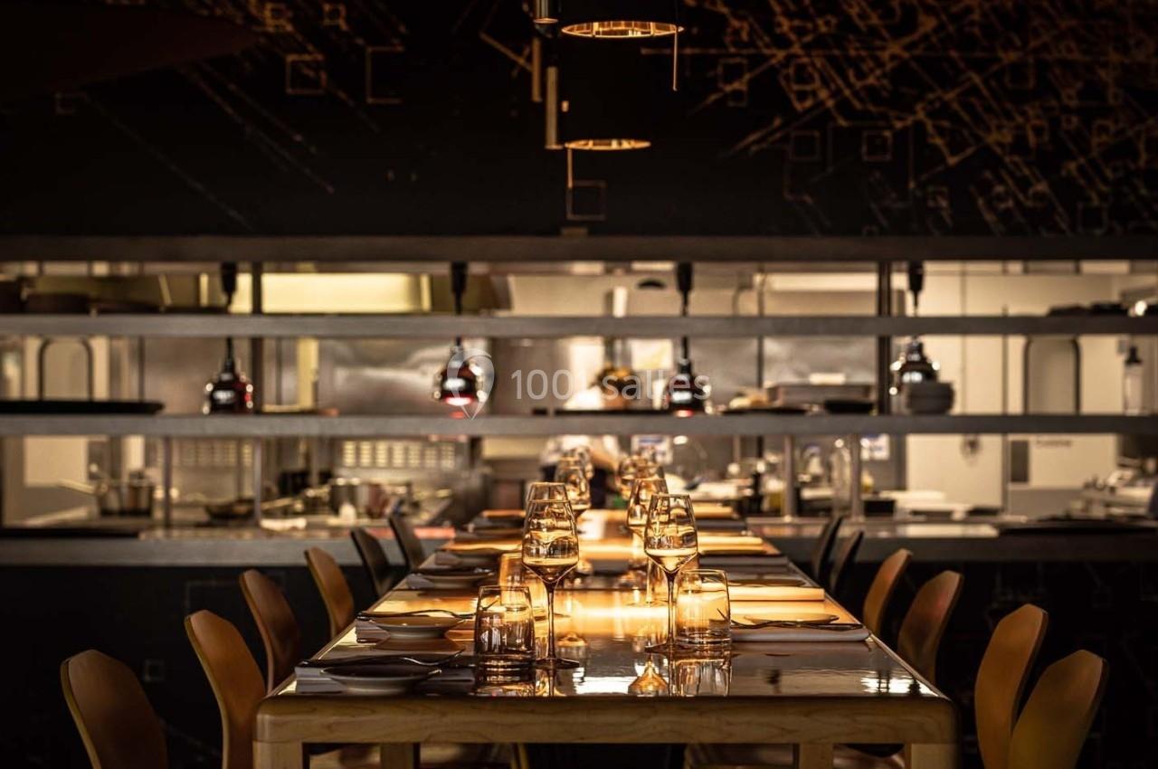 Salle de restaurant avec une table dressée, éclairée par des suspensions, et une cuisine ouverte en arrière-plan.