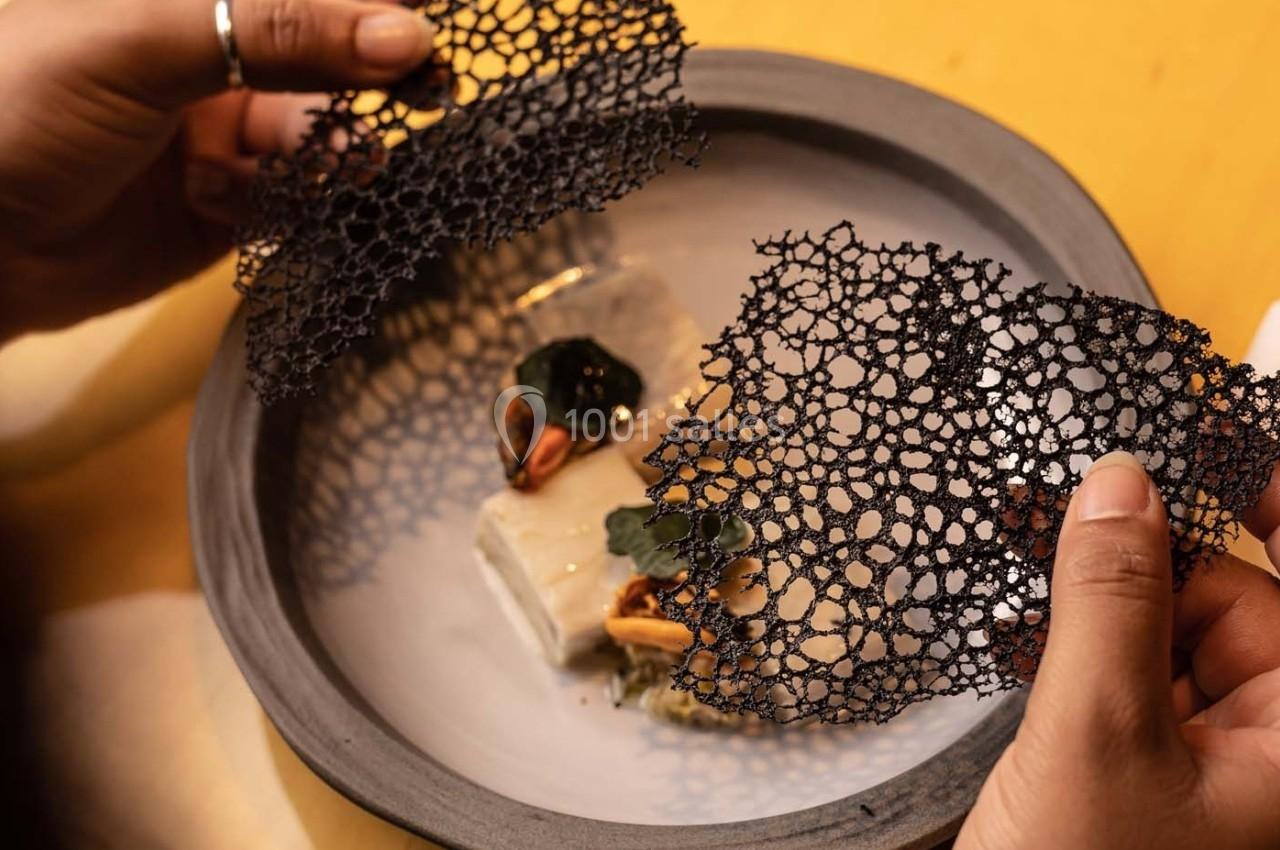 Deux mains tiennent une fine tuile noire au-dessus d'un plat gastronomique avec des éléments décoratifs.