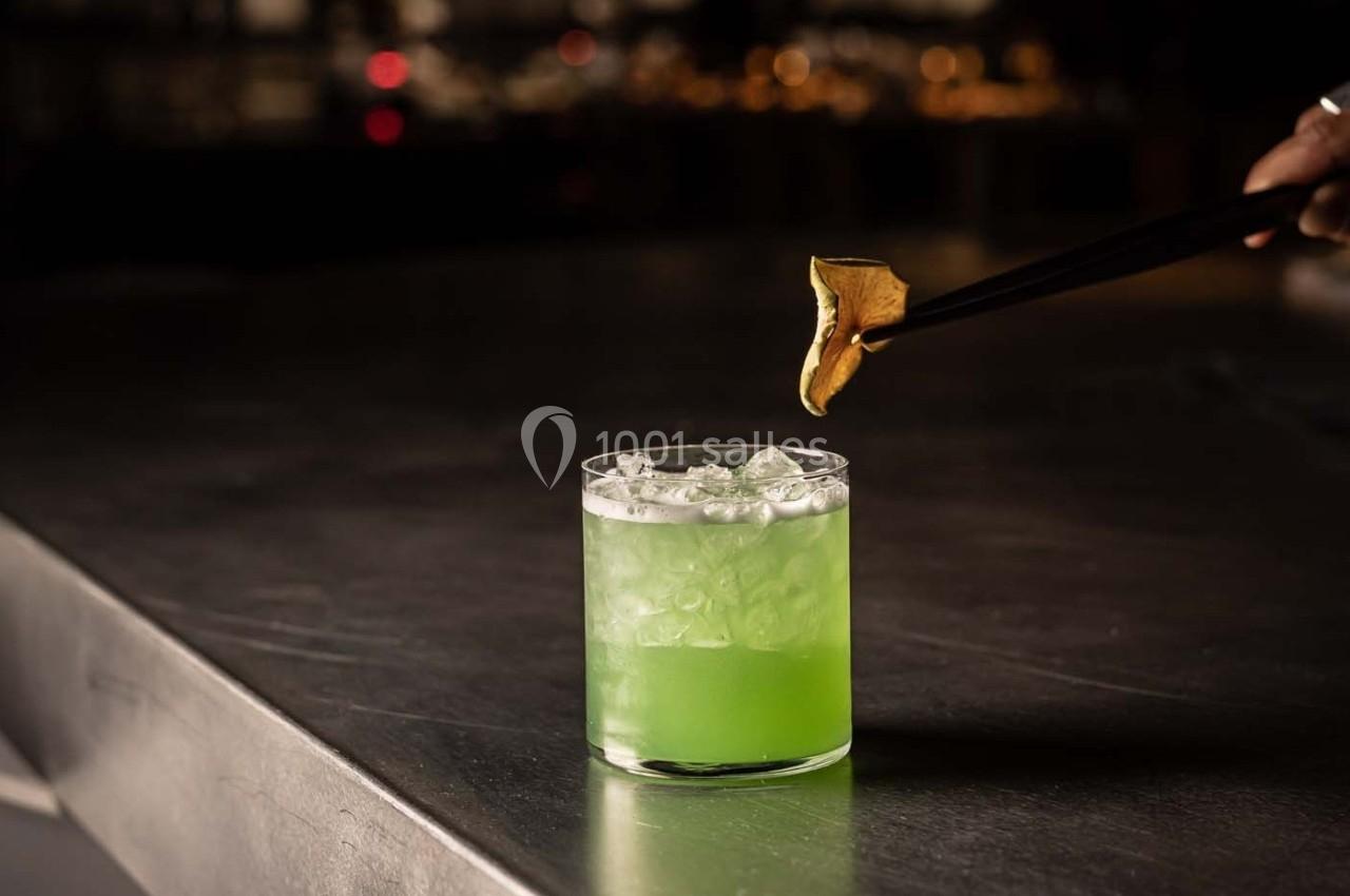 Verre de cocktail vert avec glaçons, décoré d'un zeste d'agrume doré tenu par des baguettes noires.