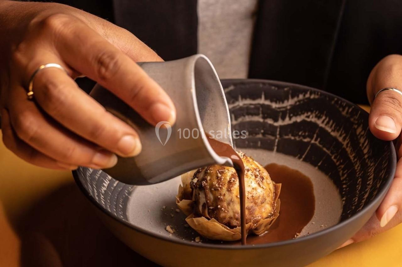 Une personne verse une sauce chocolatée sur un dessert à base de glace et de pâte croustillante dans un bol sombre.