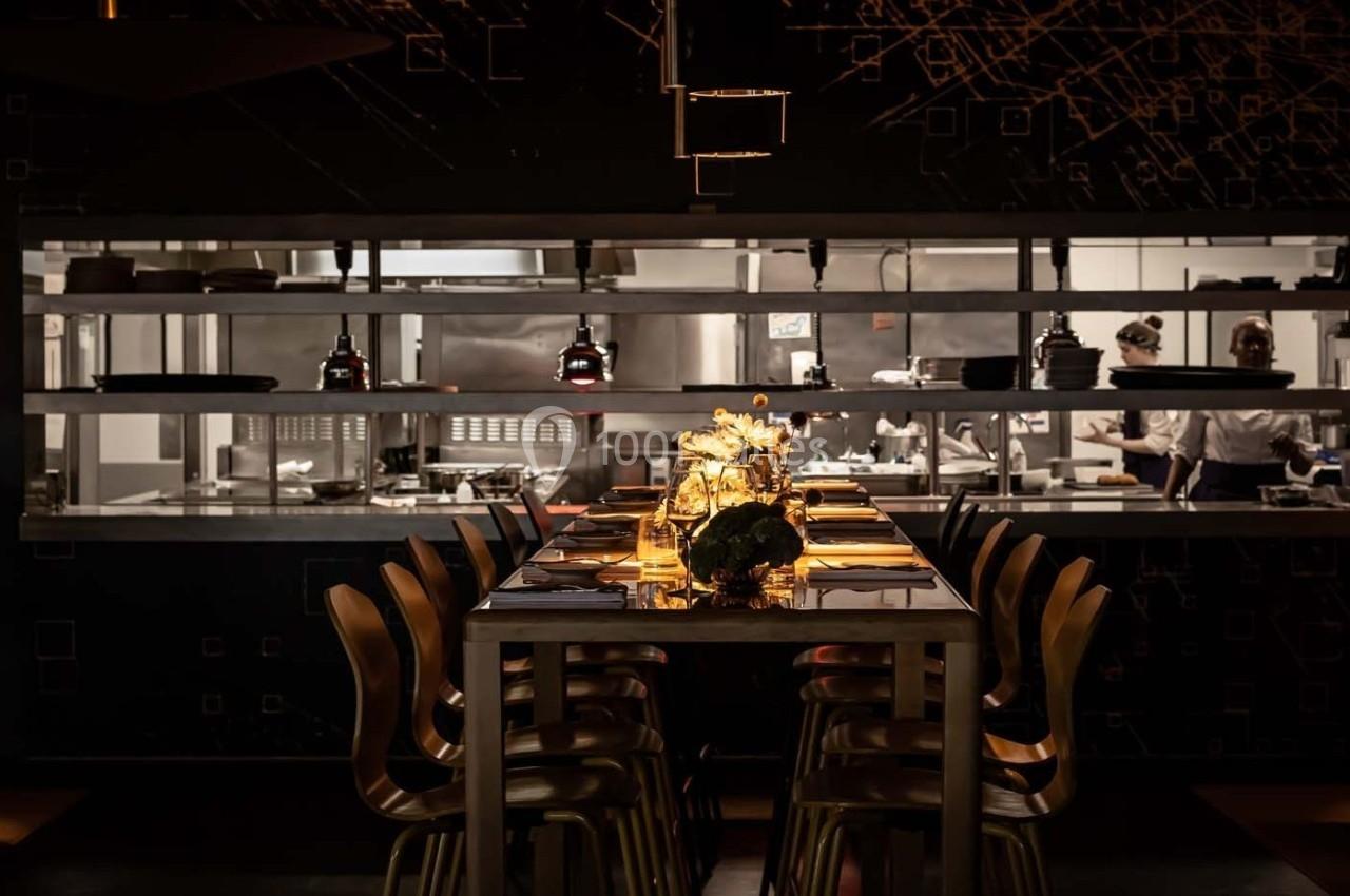 Salle de restaurant sombre avec une table dressée et éclairée, cuisine ouverte en arrière-plan avec deux chefs.